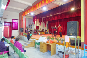 Eine Bühne mit zwei singenden Personen, eine Band im Hintergrund, rote Vorhänge, ein Licht, ein Lautsprecher rechts und ein Publikum auf Stühlen sitzend.