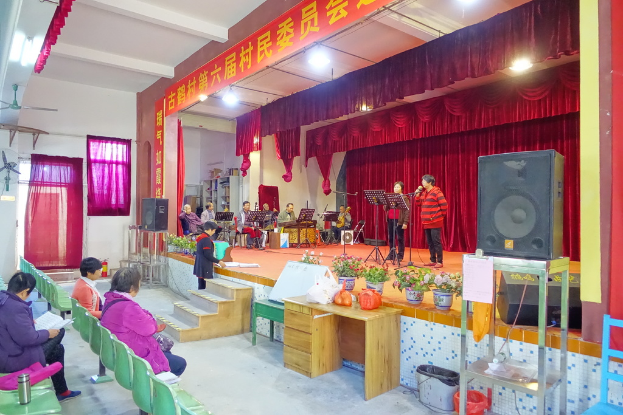 Eine Bühne mit zwei singenden Personen, eine Band im Hintergrund, rote Vorhänge, ein Licht, ein Lautsprecher rechts und ein Publikum auf Stühlen sitzend.