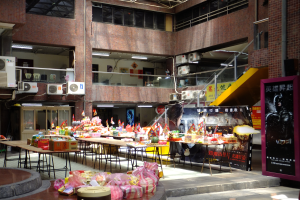 Ein geräumiger Speiseraum mit zahlreichen Tischen und Stühlen, verschiedenen Speisen auf dem Tisch, wandmontierten Klimaanlagen, Deckenleuchten und einer Tafel mit Schrift.