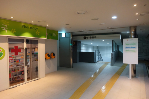 Eingang einer Apotheke mit einem "Apotheke"-Schild auf der linken Seite, einer Tafel mit Text auf der rechten Seite, beleuchteten Lampen oben und sichtbaren Gegenständen im Hintergrund.