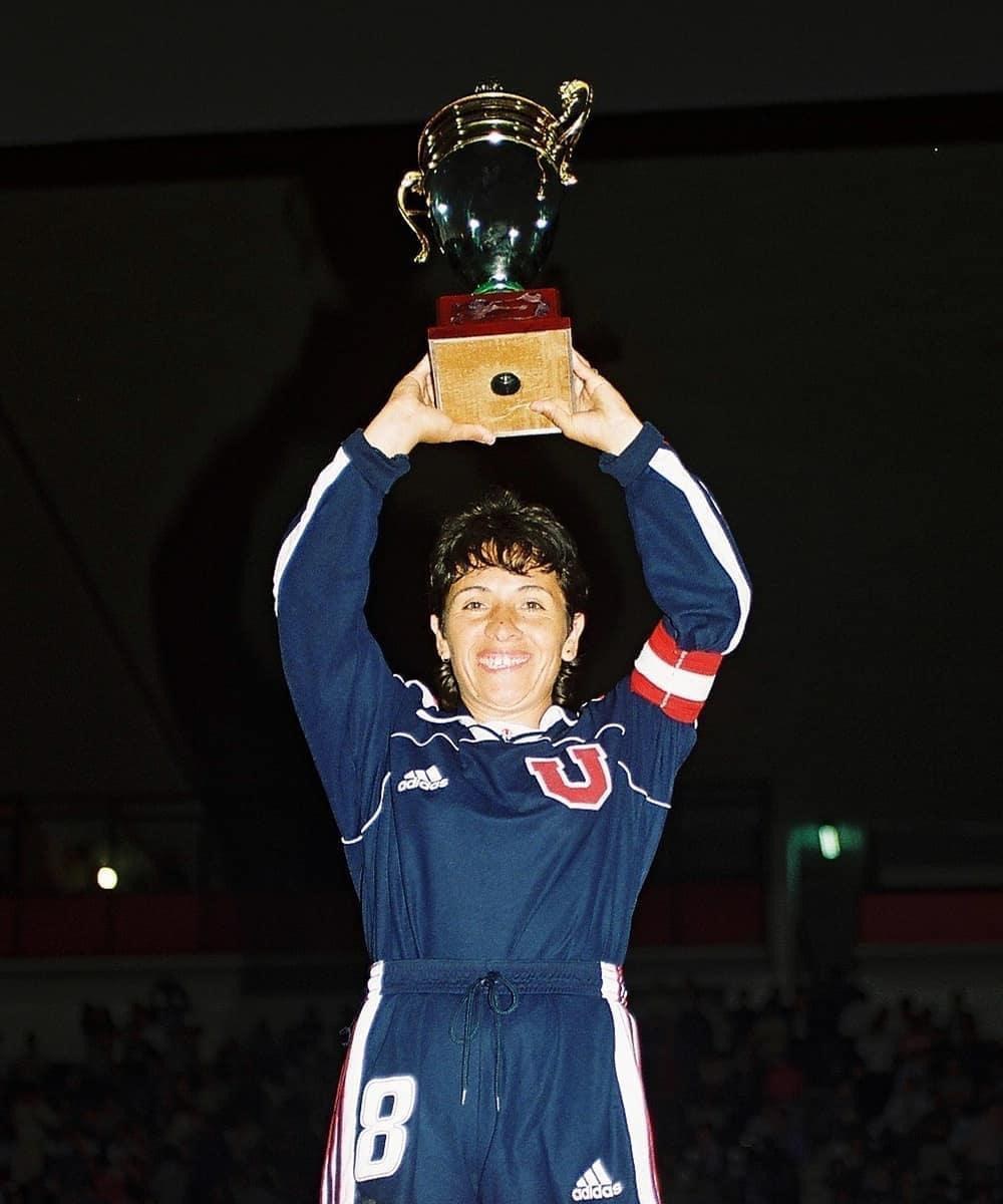 Eine Frau in einem blauen Kleid hält eine Trophäe hoch und strahlt, umgeben von einer jubelnden Menge in einem hell erleuchteten Stadion.