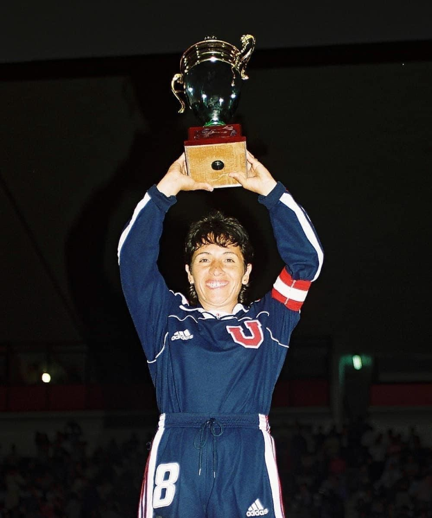 Eine Frau in einem blauen Kleid hält eine Trophäe hoch und strahlt, umgeben von einer jubelnden Menge in einem hell erleuchteten Stadion.
