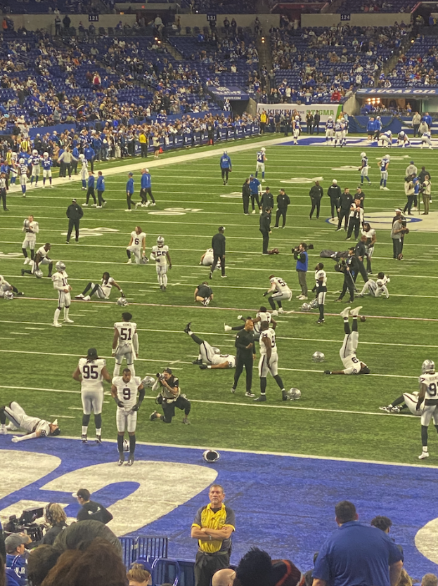 Ein Fußballspiel wird in einem Stadion gespielt, während Menschen auf dem Boden stehen und einige auf Stühlen sitzen. Eine Person hält eine Kamera, was auf eine Live-Übertragung hinweist.