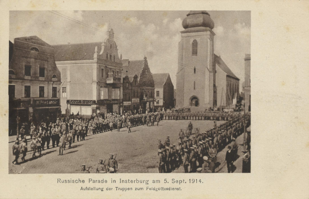 Ein Schwarz-Weiß-Foto eines Umzugs in Insterburg am 5. September 1914 mit vielen Menschen, Gebäuden und einem bewölkten Himmel sowie einem Text am unteren Rand.