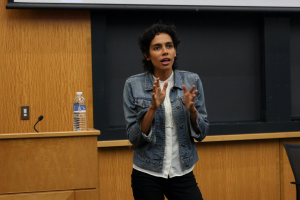 Eine Frau steht an einem Rednerpult mit einem Mikrofon und einer Flasche, hält eine Präsentation vor einem Projektionsschirm, mit einem Stuhl rechts und einer hölzernen Wand im Hintergrund.