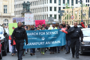 Eine Gruppe von Menschen marschiert auf einer Straße und hält ein "March for Science Frankfurt am Main"-Schild, mit Autos, Gebäuden, Statuen, Laternenmasten, Schildern und Bäumen im Hintergrund.