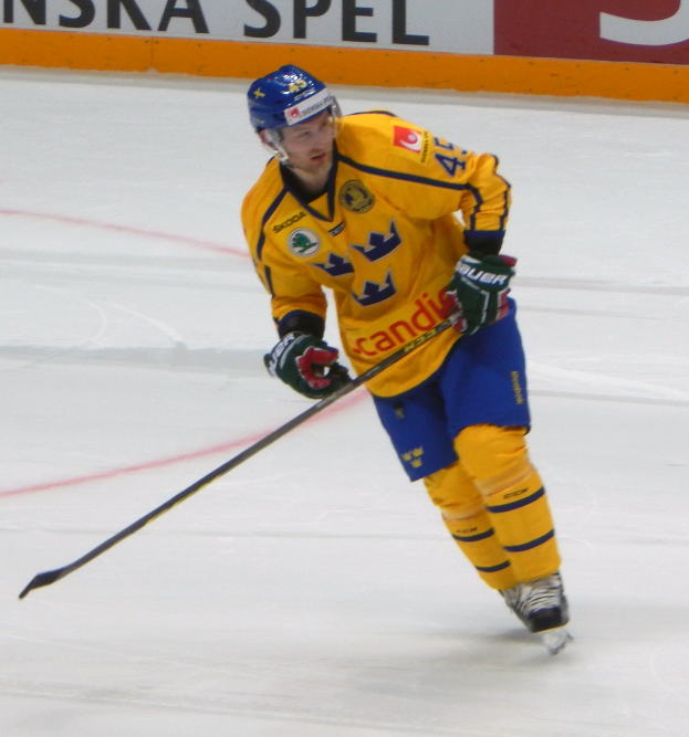 Ein Eishockeyspieler in Aktion auf dem Eis, der einen Helm, Handschuhe und Schlittschuhe trägt und einen Eishockeyschläger hält, mit einer Tafel mit Text im Hintergrund.