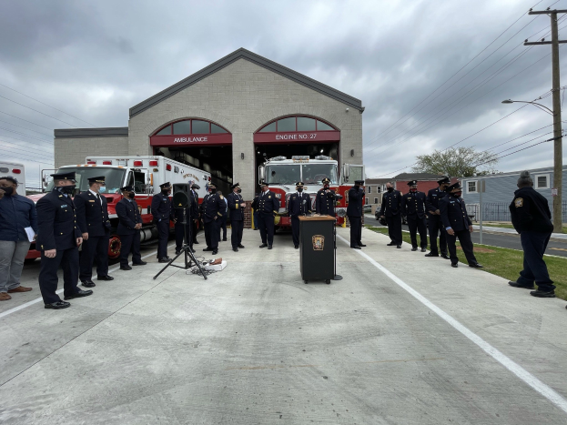 Gruppe von Menschen in Uniformen vor einem Löschfahrzeug auf einem Podium während einer Feuerwache-Einweihung, mit Gebäuden, Bäumen, Strommasten und einem bewölkten Himmel im Hintergrund.