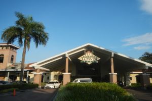 Der Eingang eines Resorts mit Palmen vorne, umgeben von Gebäuden, Kraftfahrzeugen auf der Straße, Verkehrskegeln, Büschen, Bäumen und einem bewölkten Himmel im Hintergrund.