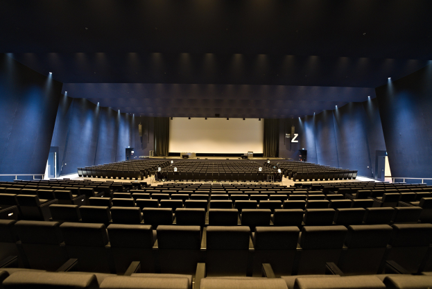 Ein großer Zuschauerraum mit Sitzreihen, einem Projektionsschirm und Bühnenobjekten, beleuchtet von Wandlampen.