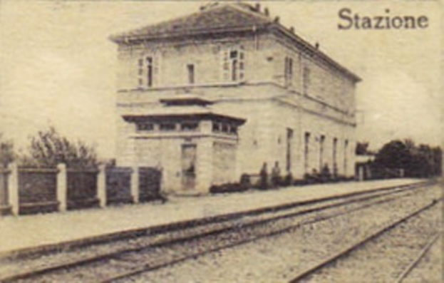Altes Schwarz-Weiß-Foto eines Bahnhofsgebäudes in der Mitte zwischen Bäumen, mit einem Zaun auf der linken Seite und Eisenbahnschienen unten; Text erscheint oben.