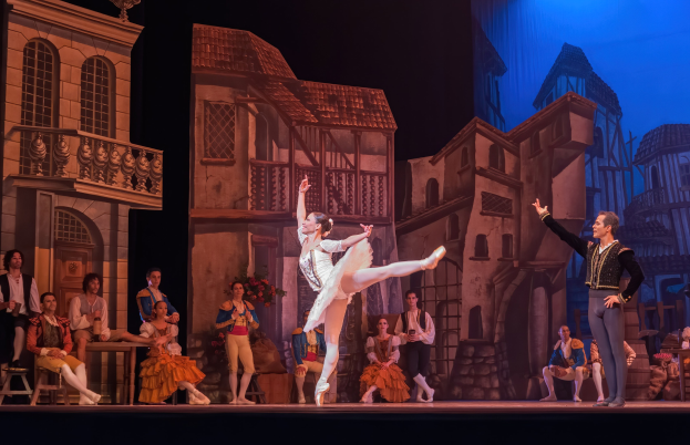 Zwei Tänzer performen auf einer Bühne im Royal Opera House in London, mit einer projizierten Kulisse von Gebäuden und einem Publikum, das sitzt und steht.