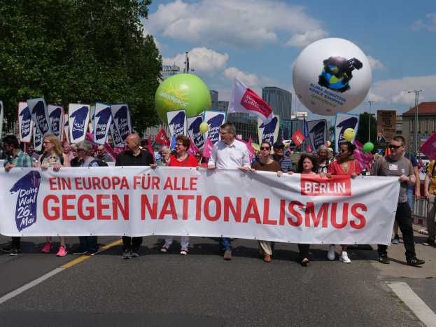 Eine Gruppe von Menschen marschiert bei einer Demonstration gegen Nationalismus in Berlin, sie halten Schilder, Fahnen und Ballons, im Hintergrund sind Bäume, Gebäude und ein bewölkter Himmel zu sehen.