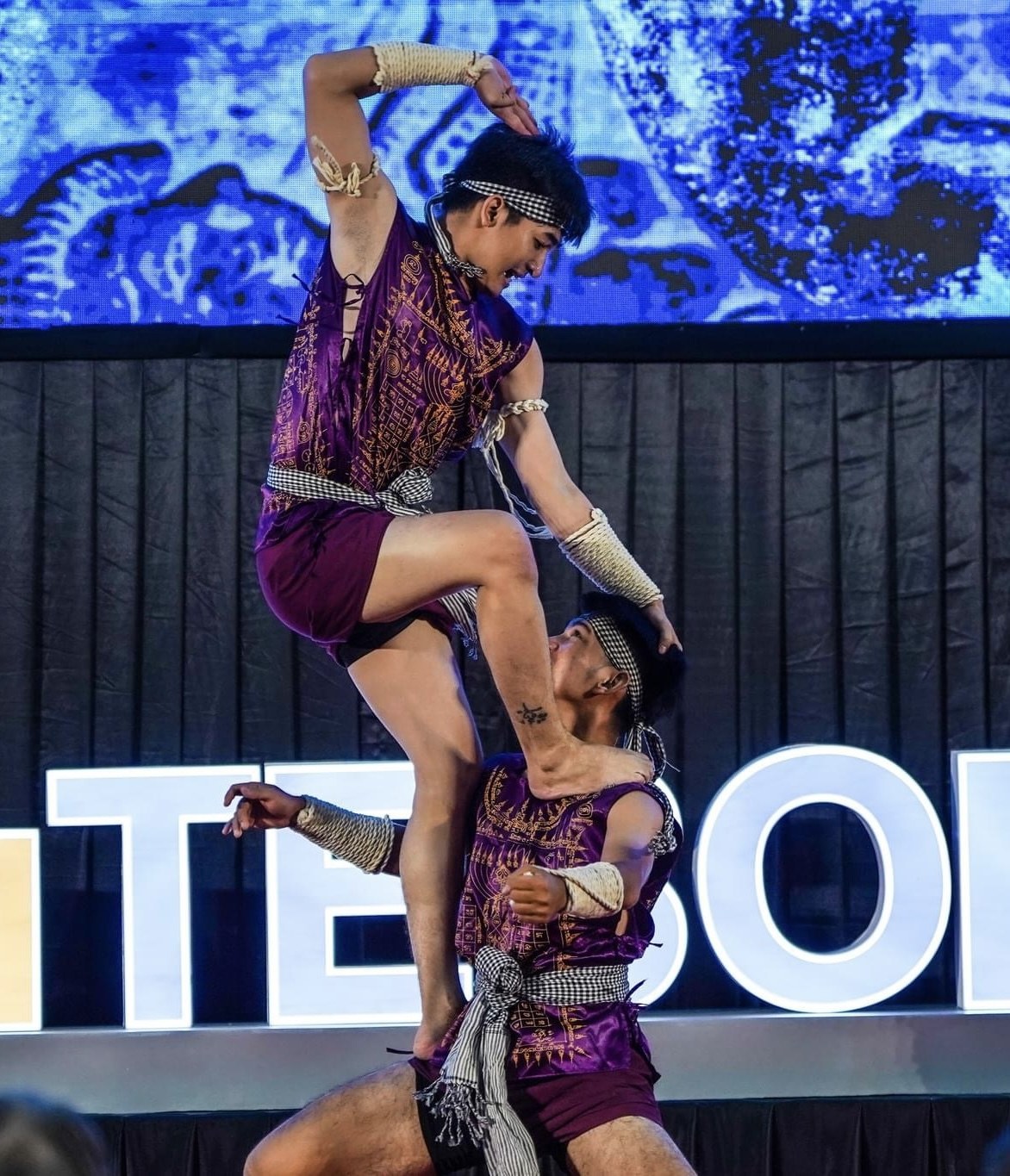 Zwei Akrobaten führen auf der Bühne eine Darbietung auf, wobei der Mann die Frau in seinen Armen hält, während beide purpurne Kleider tragen, mit einem schwarzen Vorhang und einer Leinwand im Hintergrund und einem kleinen Publikum darunter.