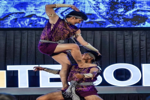 Zwei Akrobaten führen auf der Bühne eine Darbietung auf, wobei der Mann die Frau in seinen Armen hält, während beide purpurne Kleider tragen, mit einem schwarzen Vorhang und einer Leinwand im Hintergrund und einem kleinen Publikum darunter.