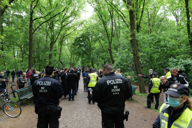Eine Gruppe von Polizisten steht vor einer Menge während einer Anti-Terror-Demonstration in Berlin, mit Fahrrädern und einer Bank im Vordergrund und Bäumen im Hintergrund.
