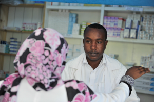 Eine Apothekerin in einem weißen Laborkittel spricht mit einer sitzenden Frau in einer Apotheke, mit Regalen voller Schachteln und anderen Gegenständen dahinter.