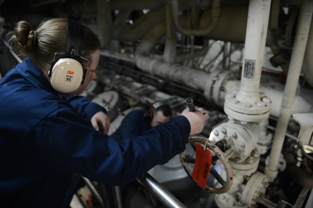 Zwei Frauen in blauen Uniformen arbeiten an einer Maschine in einer Öl- und Gasindustrieumgebung, eine hält ein Objekt und trägt Kopfhörer, mit sichtbaren Rohren im Hintergrund.