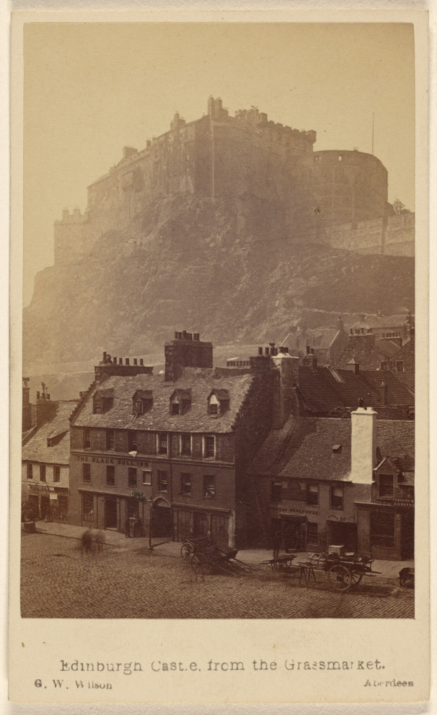 Altes Foto von Edinburgh Castle von der Grassmarket aus betrachtet, mit Gebäuden und Pferdewagen im Vordergrund und einem Hügel im Hintergrund; Text unten.
