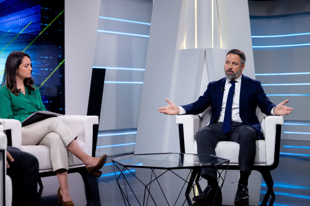 Zwei Personen sitzen auf einem TV-Set, der Mann trägt ein blaues Sakko und ein weißes Hemd mit einer blauen Krawatte, die Frau hält ein Buch in der Hand, mit einem Glas auf dem Tisch und Studiobeleuchtung im Hintergrund.