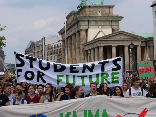Eine Gruppe von Schülern marschiert in Berlin mit einer bunten 'Students for Future'-Schleife im Hintergrund von Gebäuden, Bäumen und Himmel.