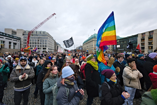 Großer Teilnehmergruppe bei einer LGBTQ+-Rechtsdemo in Berlin, mit Fahnen und Plakaten, vor einem Gebäude mit Kränen und bewölktem Himmel.