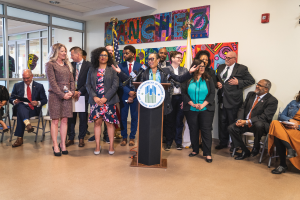 Eine Gruppe von Menschen, die vor einem Podium mit einem Mikrofon und einer Tafel mit der Aufschrift "Sancho-Stadtrat tritt dem Stadtrat bei" steht und sitzt, mit Flaggen, Feuerlöscher und Deckenleuchten im Hintergrund.
