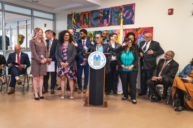 Eine Gruppe von Menschen, die vor einem Podium mit einem Mikrofon und einer Tafel mit der Aufschrift "Sancho-Stadtrat tritt dem Stadtrat bei" steht und sitzt, mit Flaggen, Feuerlöscher und Deckenleuchten im Hintergrund.