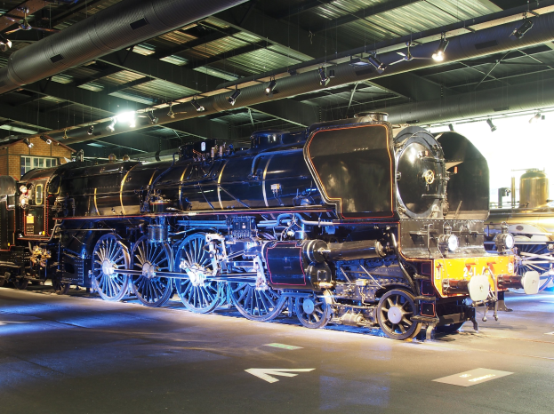 Eine große, alte Dampflokomotive, die in einem Museum ausgestellt ist, umgeben von anderen Zügen, mit einem Gebäude mit Fenstern, Lampen und einem Rohr auf dem Dach links sichtbar.