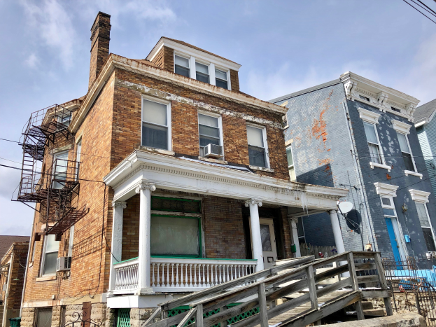 Eine Reihe baufälliger Häuser an einer Straßenecke mit zerbrochenen Fenstern, bröckelnden Wänden, Geländern, Stufen, einem Zaun, einer Satellitenschüssel, Kabeln und einem bewölkten Himmel.