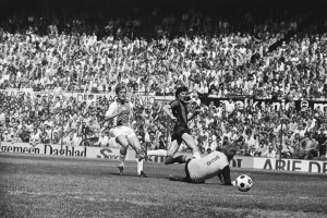 Schwarzes und weißes Foto von Männern, die auf einem Feld Fußball spielen, mit Zuschauern im Hintergrund und einem Banner "Bundesliga 1978-79 Ajax 1-1 Hannover 96"