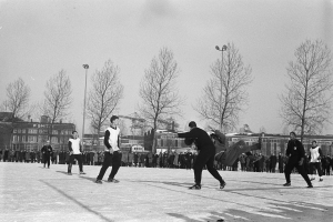 Eine Gruppe von Menschen, die im Schnee Eishockey spielen, mit Bäumen, Gebäuden, Laternen und einem klaren Himmel im Hintergrund, dargestellt in Schwarz-Weiß.