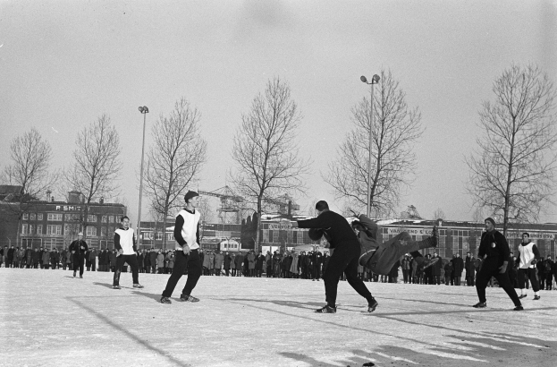Eine Gruppe von Menschen, die im Schnee Eishockey spielen, mit Bäumen, Gebäuden, Laternen und einem klaren Himmel im Hintergrund, dargestellt in Schwarz-Weiß.