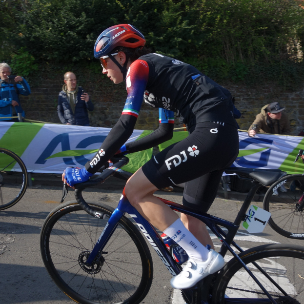 Professionelle Radsportlerin Katie Archibald mit Helm, Brille, Handschuhen und Schuhen fährt auf einem Fahrrad eine Straße entlang neben einer Menge, mit einem Banner, einer Wand, Bäumen und einem Gebäude im Hintergrund.