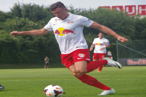 Ein professioneller Fußballspieler tritt einen Ball auf einem Rasenfeld mit "RB Leipzig" im Hintergrund unter einem klaren blauen Himmel.