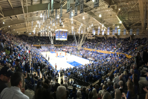 Eine große Menschenmenge, darunter Männer und Frauen, schaut ein Basketballspiel in einer Arena. Einige sitzen auf Stühlen, andere stehen auf dem Boden, während ein Bildschirm, Banner, Lichter und Stangen im Hintergrund zu sehen sind.