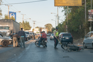 Eine Gruppe von Menschen umringt ein verunglücktes Motorrad am Stra√enrand mit mehreren Fahrzeugen, darunter ein Lastwagen, und einer Hintergrund von Bäumen, Masten, Lichtern und Schildern unter dem Himmel.