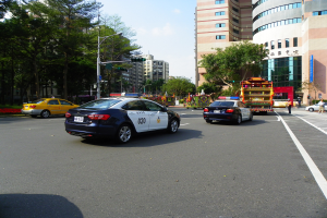 Polizeiauto fährt auf einer städtischen Straße mit hohen Gebäuden, Bäumen, Laternen, Verkehrszeichen und Passanten unter einem bewölkten Himmel.