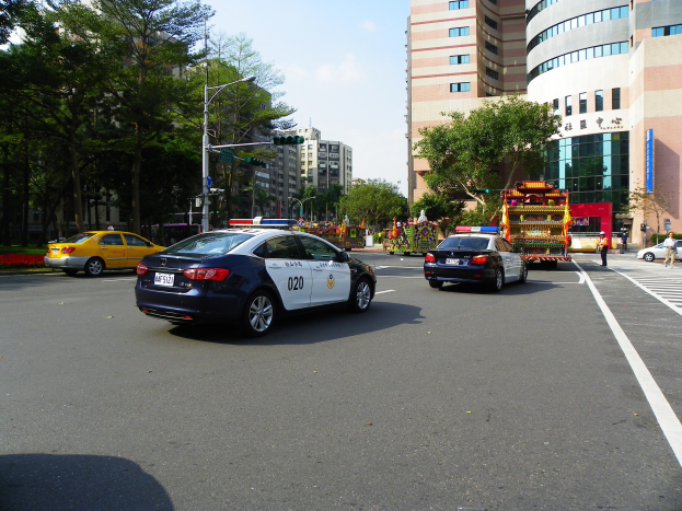 Polizeiauto fährt auf einer städtischen Straße mit hohen Gebäuden, Bäumen, Laternen, Verkehrszeichen und Passanten unter einem bewölkten Himmel.
