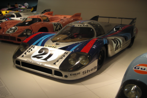 Ein Porsche 917K Le Mans Rennauto wird in einem Museum ausgestellt, umgeben von anderen Sportwagen und Menschen, mit einer Wand im Hintergrund und einem Papier mit Text auf der linken Seite.