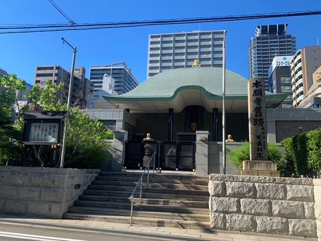 Großes grünoffenes Gebäude in einer städtischen Umgebung mit umliegenden Treppen, Geländern, Beschilderung, Grünflächen und einer Straße im Vordergrund, mit Hochhäusern und einem klaren Himmel im Hintergrund.