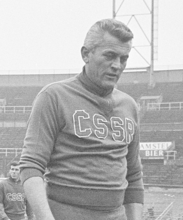 Schwarzes und weißes Foto eines Mannes, der auf einem Feld steht und ein T-Shirt mit Text trägt, mit einem Stadion, einer Werbetafel, einem Pfahl und dem Himmel im Hintergrund.