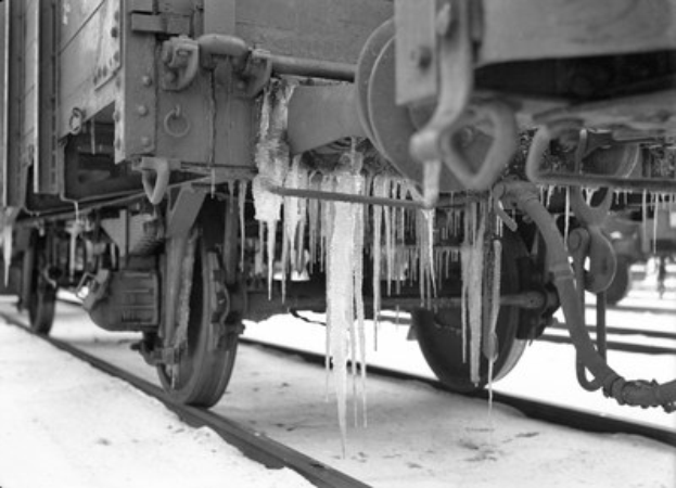 Ein Schwarz-Weiß-Foto eines Zuges auf schneebedeckten Gleisen mit Eiszapfen an seiner Seite und sichtbaren Rädern.