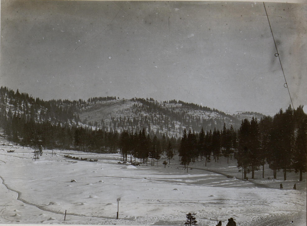 Ein Schwarz-Weiß-Foto einer Skipiste mit Menschen, die darauf Ski fahren, umgeben von Bäumen und Pfosten, mit Hügeln und einem klaren Himmel im Hintergrund und Text am unteren Rand des Bildes.