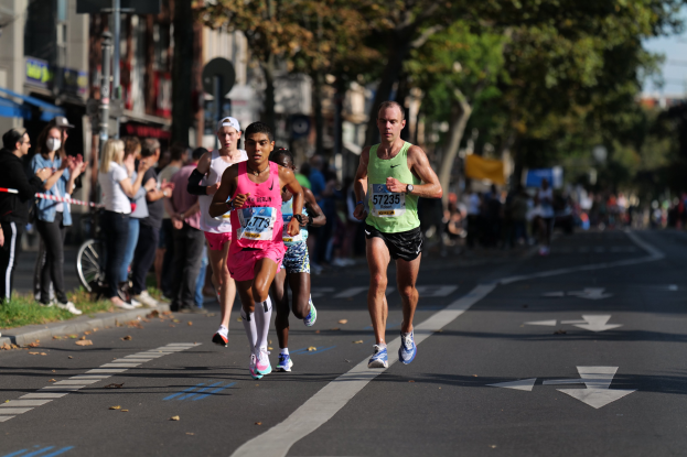 Gruppe von Menschen beim Marathon auf einer Stadtstraße mit Zuschauern, Grünflächen, Gebäuden und einem Fahrrad im Hintergrund, alles leicht unscharf.