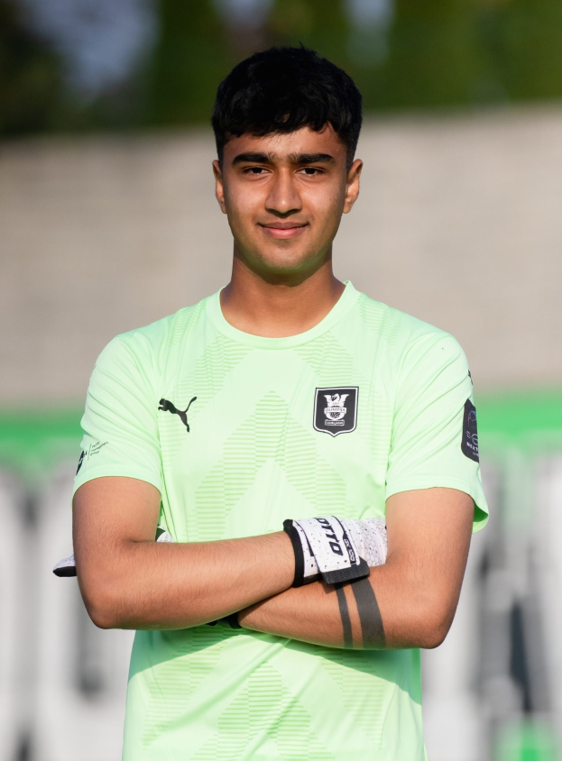 Ein junger Mann in einem grünen Hemd steht auf einem Fußballfeld und hat die Arme verschränkt, er lächelt, wahrscheinlich ein Torwart, mit einem unscharfen Hintergrund.