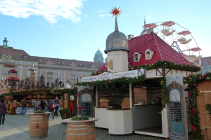 Ein geschäftiges Weihnachtsmarkt in Nürnberg, Deutschland, mit Menschen um dekorierte Stände, festliche Lichter, ein Riesenrad im Hintergrund und eine Tafel mit Text auf der rechten Seite.
