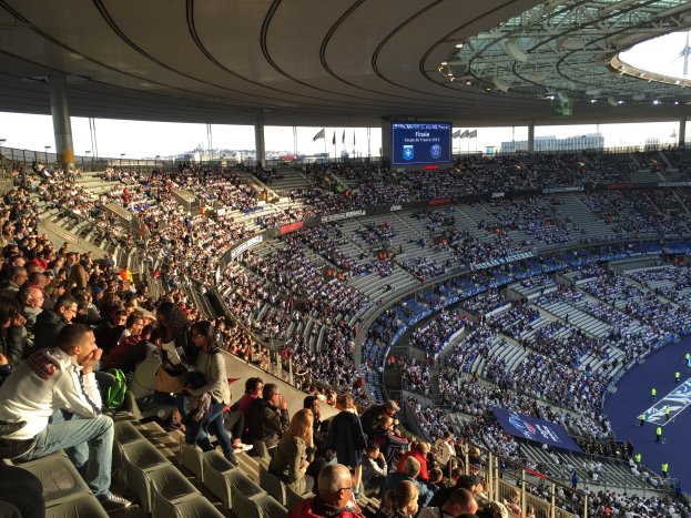 Große Menschenmenge in einem Stadion bei einem Fußballspiel mit einer Bühne, Fahnen, Stangen und einem Bildschirm im Hintergrund.