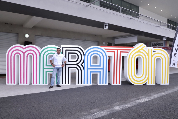 Ein Mann steht am Start eines Marathonlaufs vor einem großen "Marathon"-Schild, mit einem Banner rechts daneben und einem Gebäude mit Lampen, Geländern und Läden im Hintergrund.