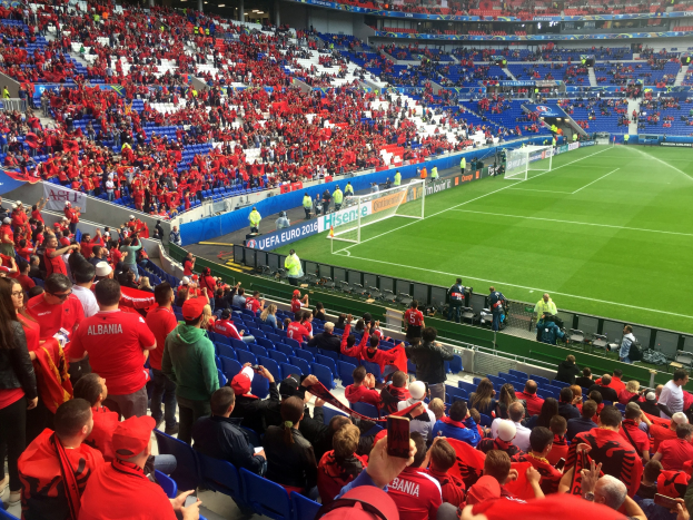 Eine große Menschenmenge in einem Stadion schaut einem Fußballspiel zu, mit Menschen auf Sitzplätzen und Stehplätzen, Bannern, einem Tor und Deckenleuchten.